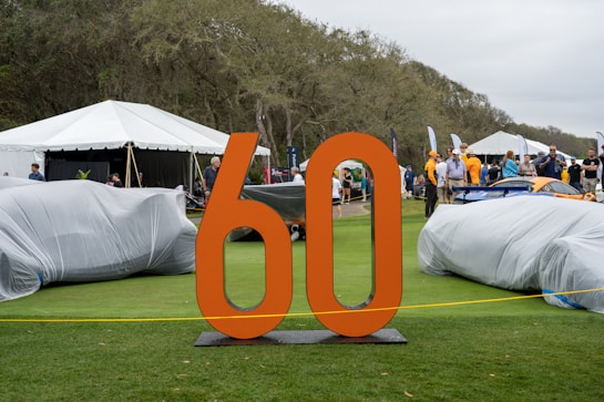 An outdoor event displays vehicles covered with protective covers on a grassy area. A large orange '60' stands prominently in the foreground. People are mingling in the background, with several tents alongside a wooded area. The setting appears casual with a mix of adults and some children.