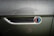 Raindrops cover the metallic surface of a vehicle, focusing on an emblem that reads 'Smit Vehicle Engineering' with a multicolored design. The background is slightly blurred, highlighting the text and colors.