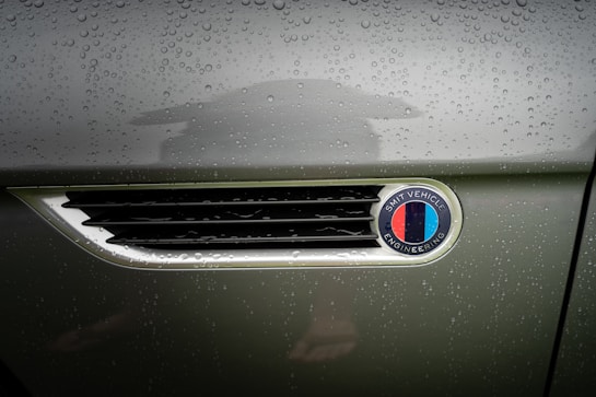 Raindrops cover the metallic surface of a vehicle, focusing on an emblem that reads 'Smit Vehicle Engineering' with a multicolored design. The background is slightly blurred, highlighting the text and colors.