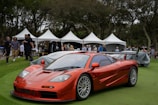 A sleek orange sports car is parked on a well-manicured lawn, likely part of a prestigious auto show or exhibition. The area is surrounded by white tents, and a group of people, including event attendees and staff, are mingling in the background. The scene is set in a park-like environment with numerous trees providing a natural backdrop.