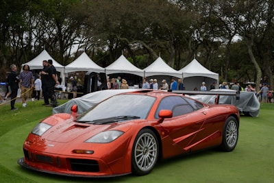 A sleek orange sports car is parked on a well-manicured lawn, likely part of a prestigious auto show or exhibition. The area is surrounded by white tents, and a group of people, including event attendees and staff, are mingling in the background. The scene is set in a park-like environment with numerous trees providing a natural backdrop.