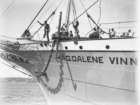 A large ship with the name 'MAGDALENE V' written on its side is shown. Several individuals are on the deck, engaged in naval activities, with one person gesturing towards something off-camera. The ship's bow is prominent, alongside large chains and an anchor. The scene occurs on a calm sea with a distant shoreline visible.