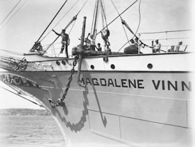 A large ship with the name 'MAGDALENE V' written on its side is shown. Several individuals are on the deck, engaged in naval activities, with one person gesturing towards something off-camera. The ship's bow is prominent, alongside large chains and an anchor. The scene occurs on a calm sea with a distant shoreline visible.