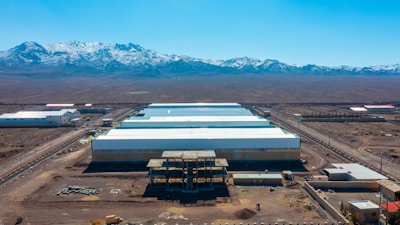 A large industrial complex with multiple white-roofed structures is situated in an arid, mountainous landscape. Snow-capped mountains rise in the background, providing a stark contrast to the dry terrain surrounding the buildings. The foreground features construction materials and partially developed structures, indicating ongoing development or expansion.