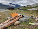 A small campfire burning with bright orange flames amongst a pile of logs and stones in a mountainous landscape. In the background, wooden cabins are visible under a cloudy sky, with mountains partially shrouded in mist. A path runs alongside a lake, adding to the serene and rustic setting.