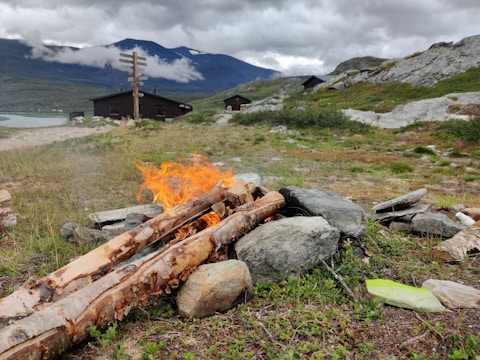 A small campfire burning with bright orange flames amongst a pile of logs and stones in a mountainous landscape. In the background, wooden cabins are visible under a cloudy sky, with mountains partially shrouded in mist. A path runs alongside a lake, adding to the serene and rustic setting.