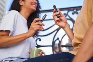 Two people are holding electronic devices, possibly vapes, in a casual setting. One person is wearing a light-colored shirt and holding a green mug. They appear to be engaged in a conversation on a balcony with ornate railing in the background.