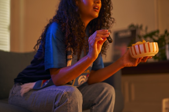 A person with curly hair is sitting on a couch holding a television remote in one hand and a bowl of snacks in the other. The person appears to be engaged in watching something on television.