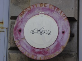 A vintage, weathered circular sign with a red border mounted on a stone wall. The white center features a simple black graphic of a tow truck pulling a car, suggesting a no-parking or tow zone. The outer edge has faded lettering, furthering the aged look.