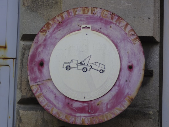 A vintage, weathered circular sign with a red border mounted on a stone wall. The white center features a simple black graphic of a tow truck pulling a car, suggesting a no-parking or tow zone. The outer edge has faded lettering, furthering the aged look.