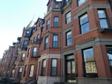 A row of historic brownstone buildings with ornate architectural details and large windows. The buildings are constructed with red brick and feature several levels, each with wrought iron balconies and black-framed windows. The sky is clear, casting soft shadows across the facades.