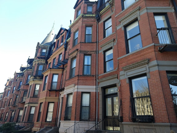 A row of historic brownstone buildings with ornate architectural details and large windows. The buildings are constructed with red brick and feature several levels, each with wrought iron balconies and black-framed windows. The sky is clear, casting soft shadows across the facades.