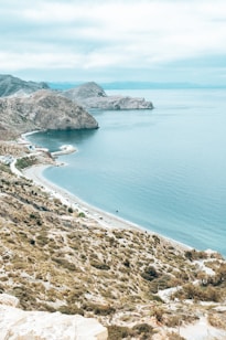 A scenic coastal landscape with rugged cliffs meeting a calm, expansive body of water under a cloudy sky. The shoreline curves elegantly with a road running parallel to it, and sparse vegetation covers the rocky terrain.