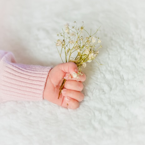 A baby's hand gently clasps a small bouquet of delicate white flowers. The background is a soft, fluffy white surface, complementing the baby's pink sweater sleeve.