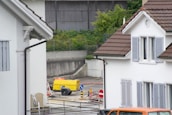 A residential area featuring two white houses with brown roofs and gray shutters. In the foreground, there is an area with roadwork signs, including a yellow portable generator and a couple of traffic barrels. Some chairs are stacked against the wall of the house on the left. There is a parked orange vehicle partially visible at the bottom right.