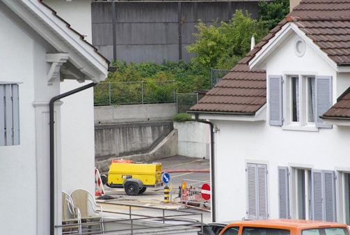 A residential area featuring two white houses with brown roofs and gray shutters. In the foreground, there is an area with roadwork signs, including a yellow portable generator and a couple of traffic barrels. Some chairs are stacked against the wall of the house on the left. There is a parked orange vehicle partially visible at the bottom right.