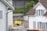 A residential area featuring two white houses with brown roofs and gray shutters. In the foreground, there is an area with roadwork signs, including a yellow portable generator and a couple of traffic barrels. Some chairs are stacked against the wall of the house on the left. There is a parked orange vehicle partially visible at the bottom right.
