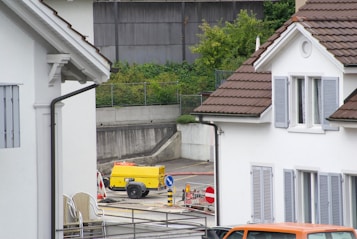 A residential area featuring two white houses with brown roofs and gray shutters. In the foreground, there is an area with roadwork signs, including a yellow portable generator and a couple of traffic barrels. Some chairs are stacked against the wall of the house on the left. There is a parked orange vehicle partially visible at the bottom right.