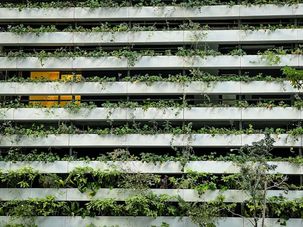 A building facade featuring horizontal rows of white concrete planters filled with lush green plants and vines. One section has a warm interior light suggesting an indoor space. The foliage varies in texture and density, giving the architecture a blend of nature and modern design.