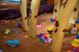 A playful indoor playground area covered in a brown, sand-like material with scattered colorful plastic toys, including toy trucks, containers, and building blocks. Large tree-like structures with a cartoonish design are situated throughout the scene, giving it a whimsical forest theme.