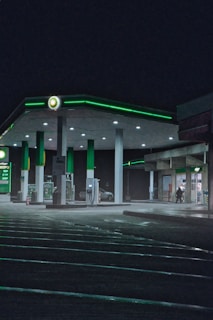 A dimly lit gas station at night features green accents and glowing logos. The station is quiet with no visible cars at the pumps. A person is entering a convenience store adjacent to the gas pumps, adding a sense of solitude to the scene. The wet ground reflects the lights, enhancing the nocturnal atmosphere.
