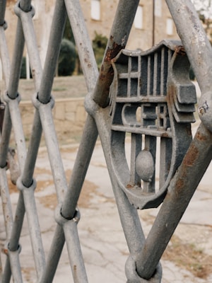 A close-up of a metal fence featuring a geometric design with a shield-like emblem at the center. The fence appears weathered with patches of rust, set against a background of blurred buildings and foliage.