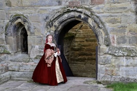 A person dressed in a historical costume stands in front of a stone archway. The attire is ornate, featuring a rich burgundy gown with intricate patterns. The stone structure behind hints at a historic or medieval setting, with weathered stones and a sense of antiquity.