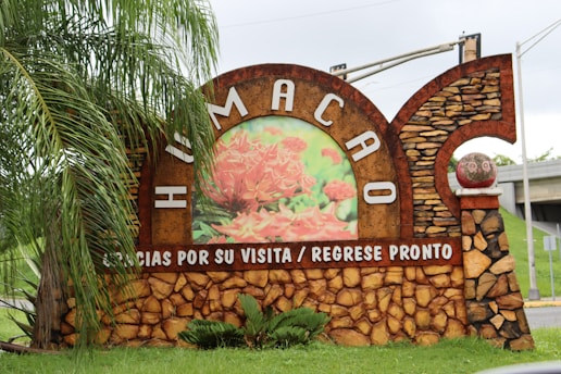 A stone sign with the word 'Humacao' in large white letters on a brown and textured background. Below it, a message reads 'Gracias por su visita / Regrese pronto' in white text on a reddish-brown band. The sign features an inset image of vibrant red flowers in the center. To the left, palm leaves partially cover the sign. To the right, a stone pillar supports a decorative red sphere. The scene is outdoors, with greenery and overcast skies in the background.