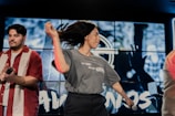A woman with long hair is energetically dancing with a microphone in her hand, while a man in a red and white striped shirt holds a microphone. The background features a large screen displaying blurred graphics or animations.