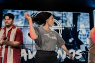 A woman with long hair is energetically dancing with a microphone in her hand, while a man in a red and white striped shirt holds a microphone. The background features a large screen displaying blurred graphics or animations.