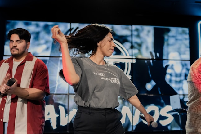 A woman with long hair is energetically dancing with a microphone in her hand, while a man in a red and white striped shirt holds a microphone. The background features a large screen displaying blurred graphics or animations.