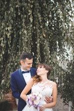 A couple, dressed in formal wedding attire, stands closely together in an outdoor setting with lush green foliage in the background. The groom is wearing a dark blue suit with a bow tie, while the bride is in a white lace wedding dress, holding a bouquet of flowers with purple and white blooms.
