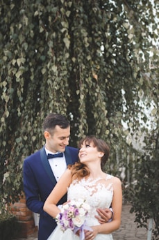 A couple, dressed in formal wedding attire, stands closely together in an outdoor setting with lush green foliage in the background. The groom is wearing a dark blue suit with a bow tie, while the bride is in a white lace wedding dress, holding a bouquet of flowers with purple and white blooms.