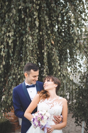 A couple, dressed in formal wedding attire, stands closely together in an outdoor setting with lush green foliage in the background. The groom is wearing a dark blue suit with a bow tie, while the bride is in a white lace wedding dress, holding a bouquet of flowers with purple and white blooms.