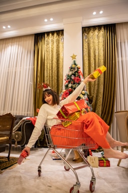A person in a white sweater and red pants sits playfully in a shopping cart, holding several brightly wrapped gifts. A decorated Christmas tree with a star on top is positioned in the background, surrounded by warm, golden curtains. The room features several chairs and a polished floor, adding to the festive atmosphere.
