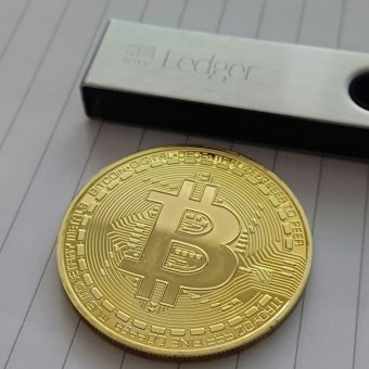 A shiny gold-colored object resembling a Bitcoin is placed on a lined surface next to a silver and black Ledger device.