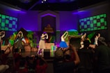 Under vibrant stage lights, a group of performers stands on stage, surrounded by colorful backdrops suggesting a natural theme with green and brown hues. They are engaged in a dance routine, with some individuals wearing costumes with bright elements like a blue tutu. An audience, visible in silhouette, watches and raises arms, creating an interactive atmosphere.