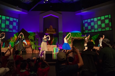 Under vibrant stage lights, a group of performers stands on stage, surrounded by colorful backdrops suggesting a natural theme with green and brown hues. They are engaged in a dance routine, with some individuals wearing costumes with bright elements like a blue tutu. An audience, visible in silhouette, watches and raises arms, creating an interactive atmosphere.