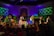 Under vibrant stage lights, a group of performers stands on stage, surrounded by colorful backdrops suggesting a natural theme with green and brown hues. They are engaged in a dance routine, with some individuals wearing costumes with bright elements like a blue tutu. An audience, visible in silhouette, watches and raises arms, creating an interactive atmosphere.