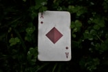 An Ace of Diamonds playing card lies on a bed of green clover leaves. The card is slightly worn and weathered, with a large diamond symbol in the center and smaller diamond icons in the corners.