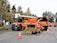 An orange maintenance truck is parked with a wood chipper working on debris. Traffic cones surround the setup on a paved street. Tall trees and a brick wall in the background add to the suburban setting.