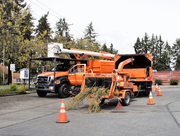 An orange maintenance truck is parked with a wood chipper working on debris. Traffic cones surround the setup on a paved street. Tall trees and a brick wall in the background add to the suburban setting.