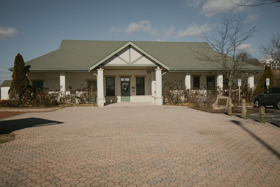 A single-story building with a light green gabled roof and a paved courtyard in front. The facade includes white walls, green trim, and a central entrance with columns. Leafless trees and shrubs surround the structure, and there is a clear blue sky with scattered clouds overhead.