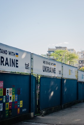 A series of fences displays banners with the phrase 'WE STAND WITH UKRAINE' alongside the Ukrainian flag and the logo 'taf'. A wall features colorful square panels with painted faces and expressions. In the background, there are buildings and a clear blue sky.