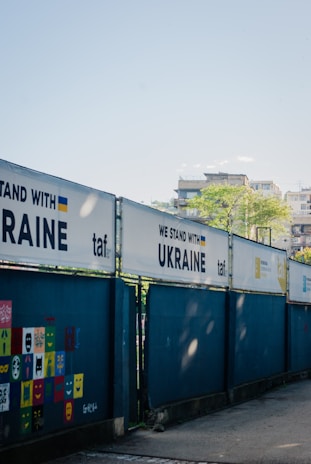 A series of fences displays banners with the phrase 'WE STAND WITH UKRAINE' alongside the Ukrainian flag and the logo 'taf'. A wall features colorful square panels with painted faces and expressions. In the background, there are buildings and a clear blue sky.