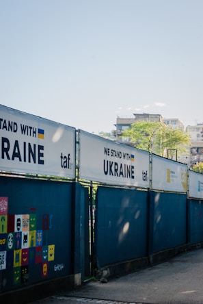 A series of fences displays banners with the phrase 'WE STAND WITH UKRAINE' alongside the Ukrainian flag and the logo 'taf'. A wall features colorful square panels with painted faces and expressions. In the background, there are buildings and a clear blue sky.