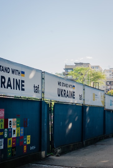 A series of fences displays banners with the phrase 'WE STAND WITH UKRAINE' alongside the Ukrainian flag and the logo 'taf'. A wall features colorful square panels with painted faces and expressions. In the background, there are buildings and a clear blue sky.