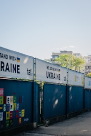A series of fences displays banners with the phrase 'WE STAND WITH UKRAINE' alongside the Ukrainian flag and the logo 'taf'. A wall features colorful square panels with painted faces and expressions. In the background, there are buildings and a clear blue sky.