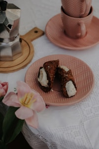 A table is set with a pink plate containing two cannoli desserts. Nearby, a metal stovetop espresso maker rests on a wooden coaster. Two matching pink cups are stacked on another pink plate. A pink flower is placed on the white lace tablecloth, adding a touch of color and elegance.