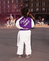 A young child, dressed in a white martial arts uniform with a purple belt, stands with their back to the camera. The purple top features an inspiring message written in Spanish. In the background, a large plaza appears with several people in various poses, suggesting a public event or gathering.